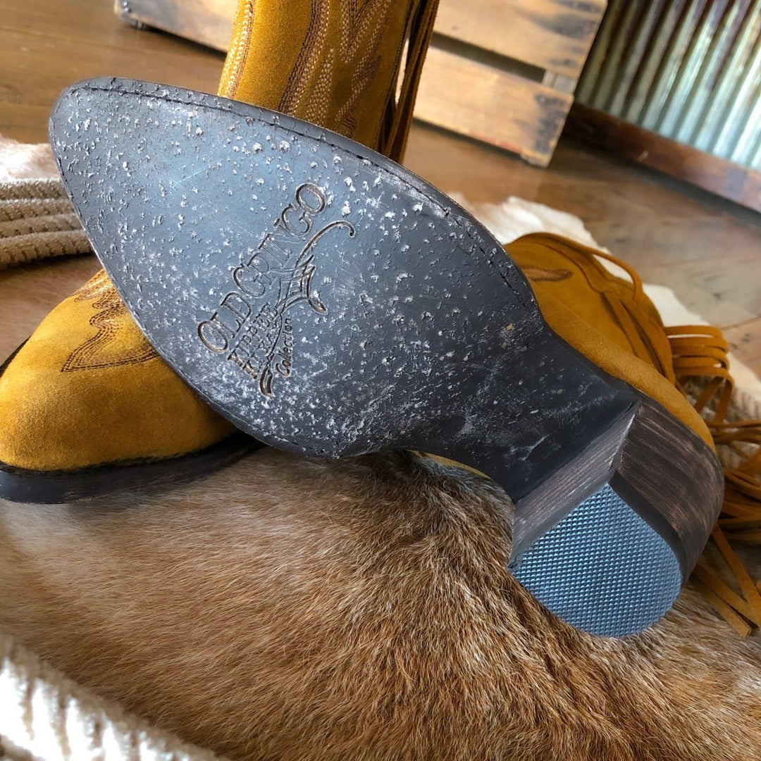 Close-up of the sole of The Tejas by Savannah Sevens western life{&}style, a yellow suede boot with decorative patterns, resting on a fur rug with another boot visible in the background.