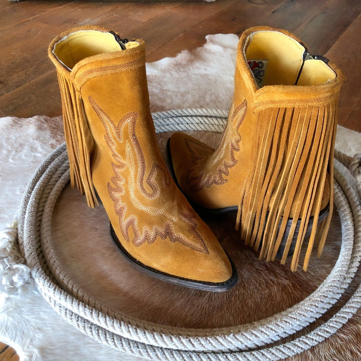 A pair of The Tejas tan suede booties by Savannah Sevens western life{&}style with leather fringe and decorative stitching sit on a cowhide rug, inside a coiled rope, on a wooden floor.