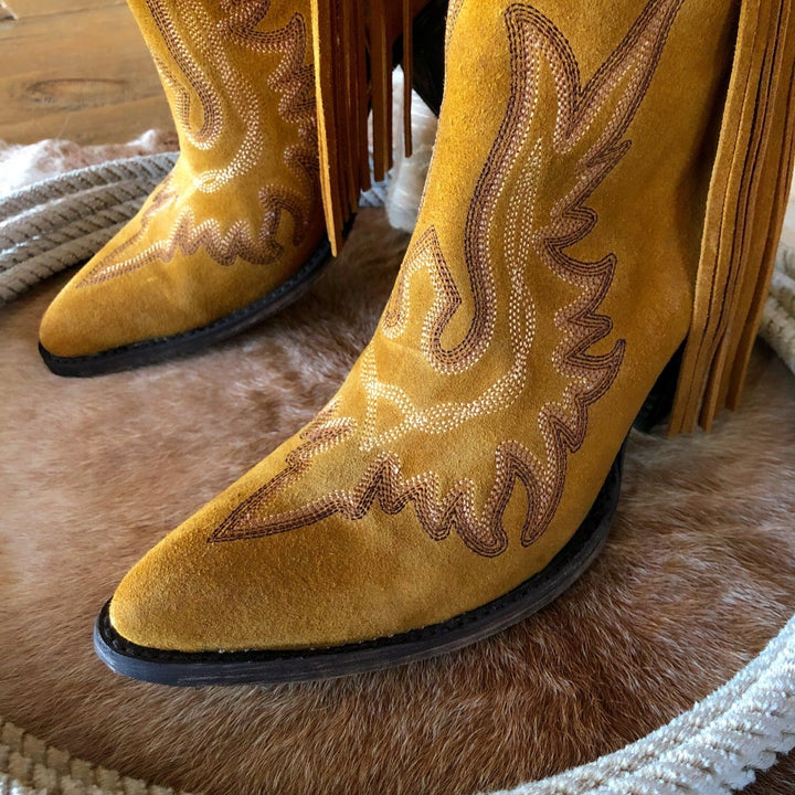 Close-up of The Tejas pointed-toe yellow suede western boots by Savannah Sevens western life{&}style, featuring decorative stitching and leather fringe, resting on animal hide beside a coiled rope.
