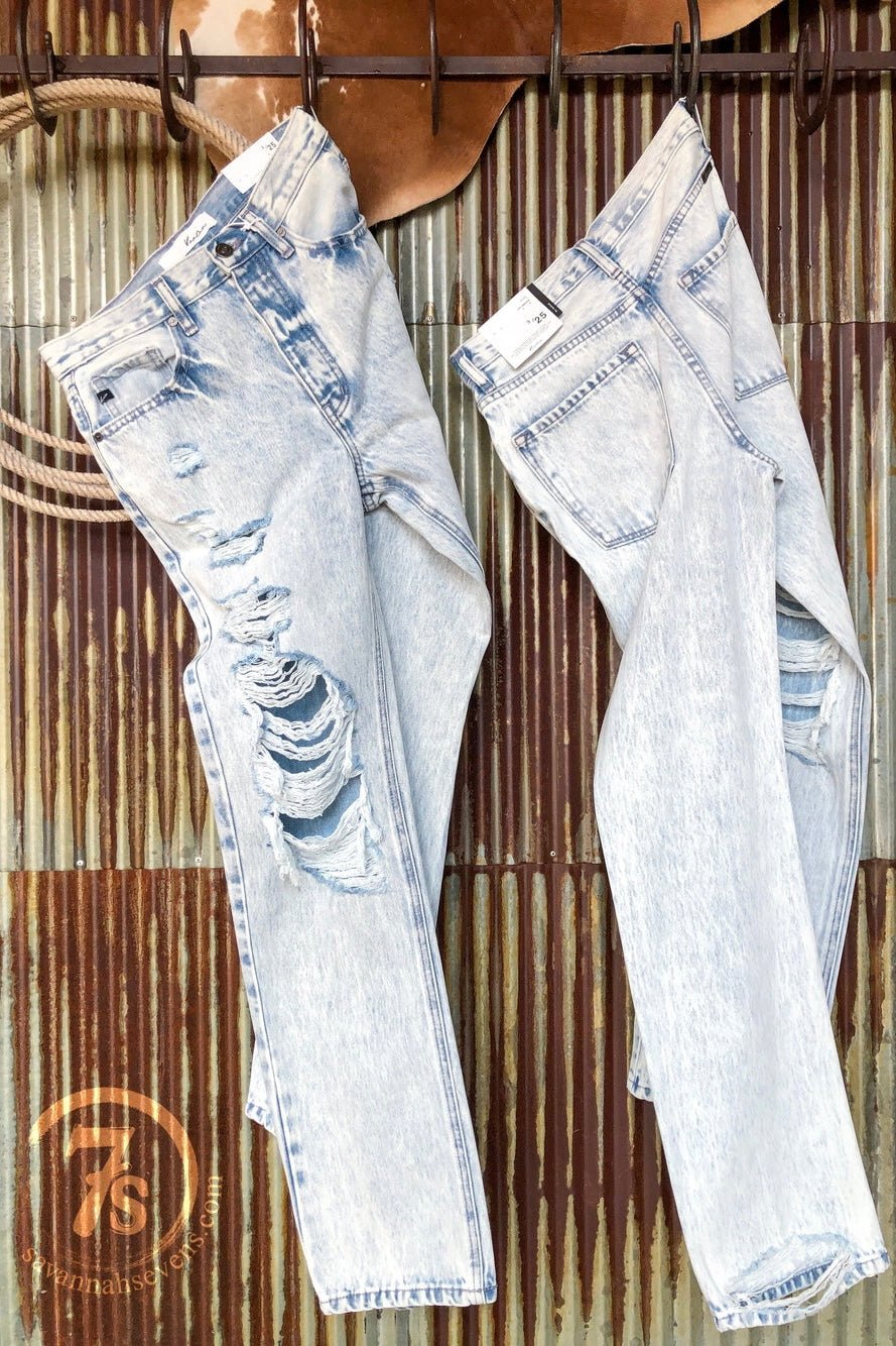 Two pairs of light-wash blue jeans hang on hooks against a corrugated metal wall: one is destroyed denim with rips; the other is The Wyatt High Rise 90's Straight Leg Jean by Savannah Sevens western life{&}style. A round 7s logo appears bottom left.