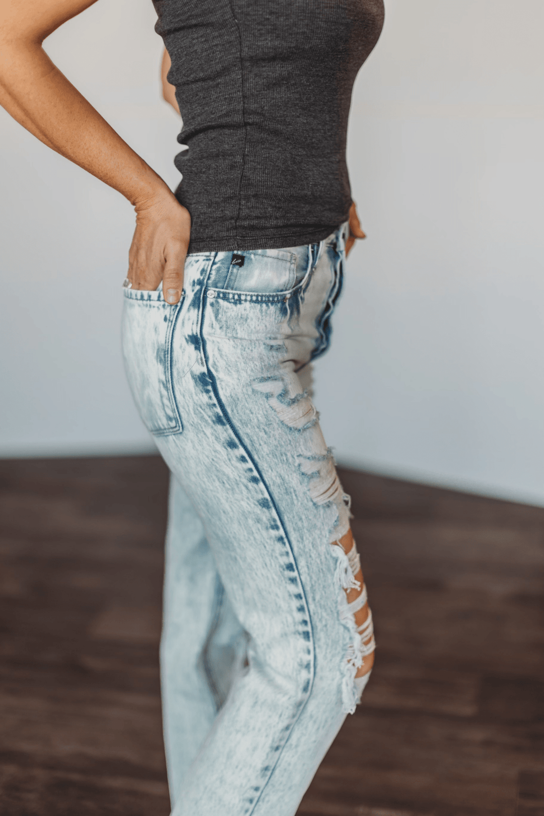 A person stands with one hand on their hip, wearing The Wyatt High Rise 90's Straight Leg Jean by Savannah Sevens western life{&}style and a dark sleeveless top, against a blurred indoor background.