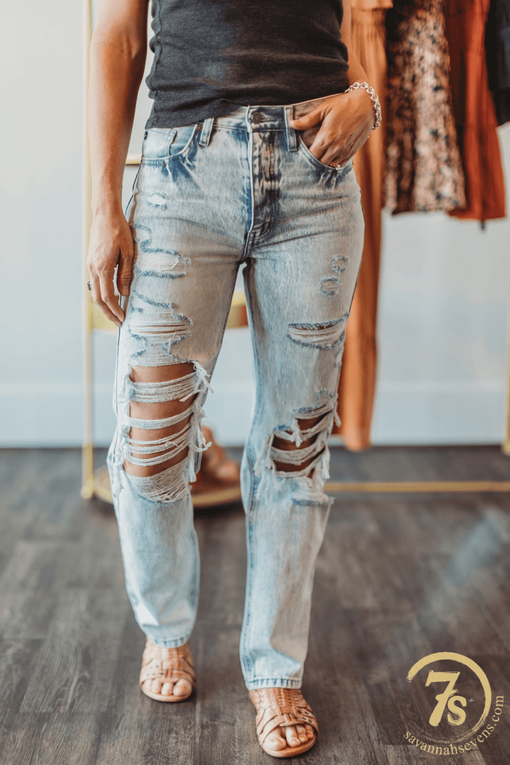 A person stands indoors wearing The Wyatt High Rise 90's Straight Leg Jean by Savannah Sevens western life{&}style, styled with a black top, tan sandals, and a bracelet. The Savannah Sevens logo is visible in the corner.