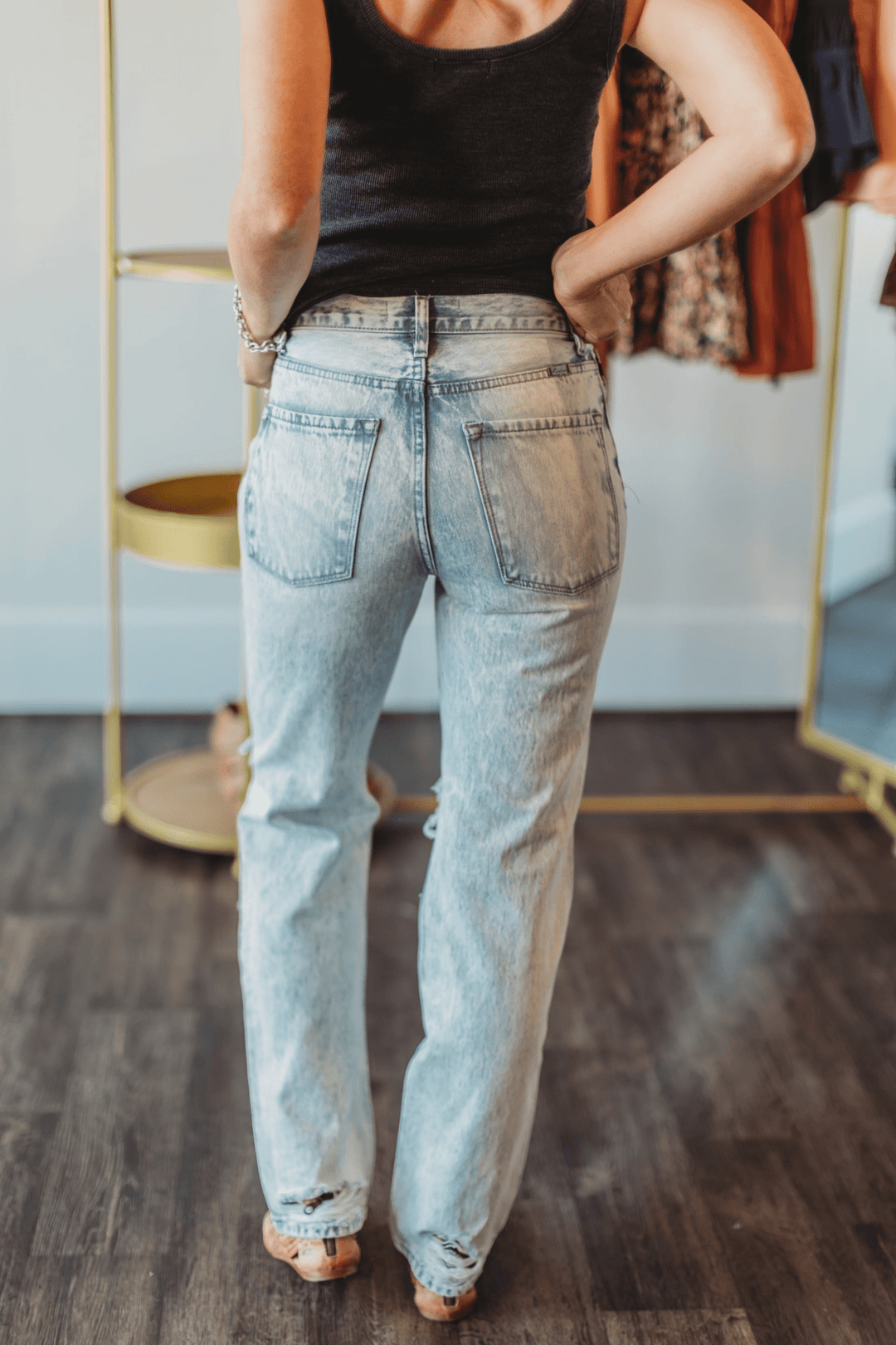 A person in a black tank top and The Wyatt High Rise 90’s Straight Leg Jean by Savannah Sevens western life{&}style stands indoors on a dark wooden floor, facing away from the camera with one hand on their hip; clothing racks are visible behind.