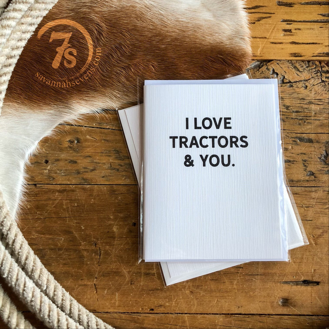 Hungry Canyon’s Tractors & You Card sits on a rustic wooden surface with a coiled rope, cowhide, and another envelope, creating a charming country-themed scene. The card reads "I LOVE TRACTORS & YOU.