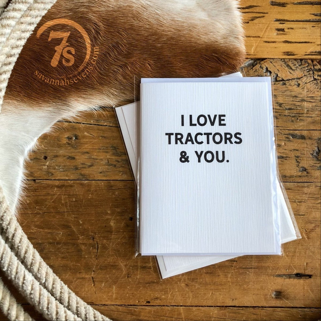 Hungry Canyon’s Tractors & You Card sits on a rustic wooden surface with a coiled rope, cowhide, and another envelope, creating a charming country-themed scene. The card reads "I LOVE TRACTORS & YOU.